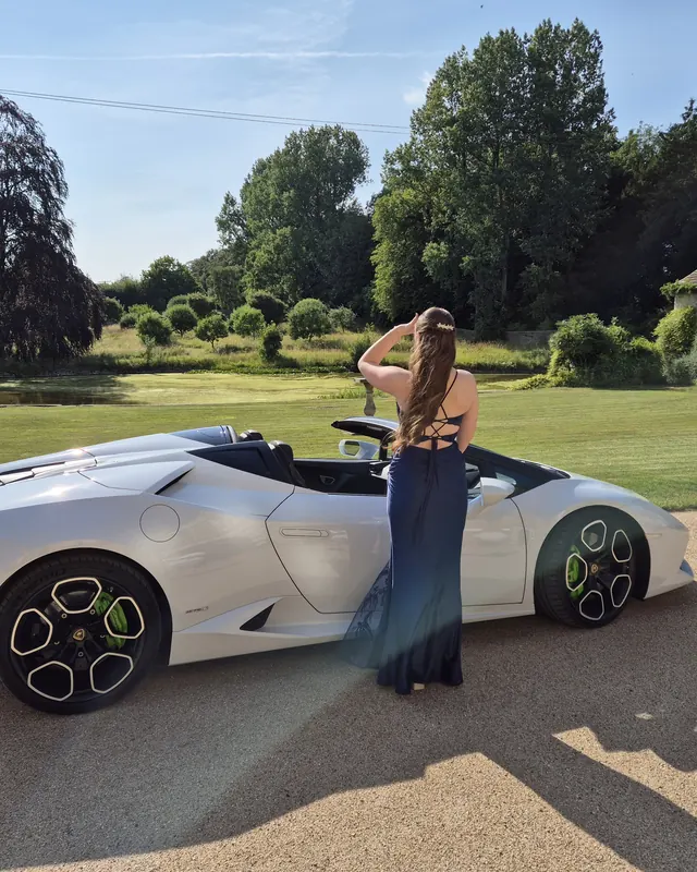 A girl in a prom dress beside a white Lamborghini at a country house