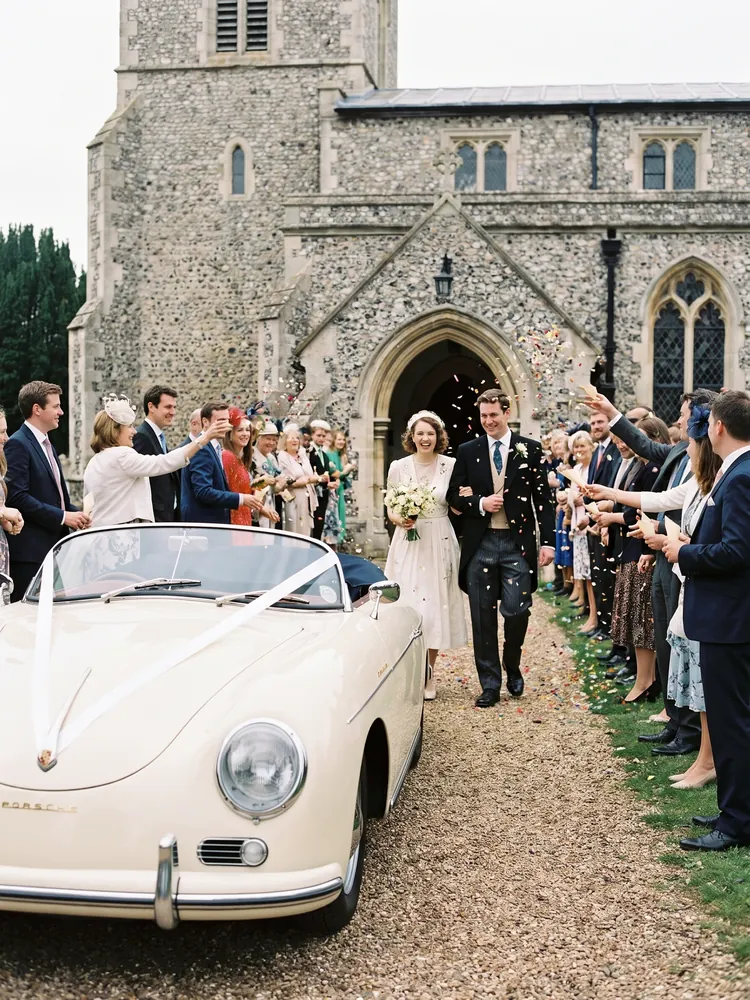 A bride and groom walking to a cream Porsche 356 Speedster outside a village church